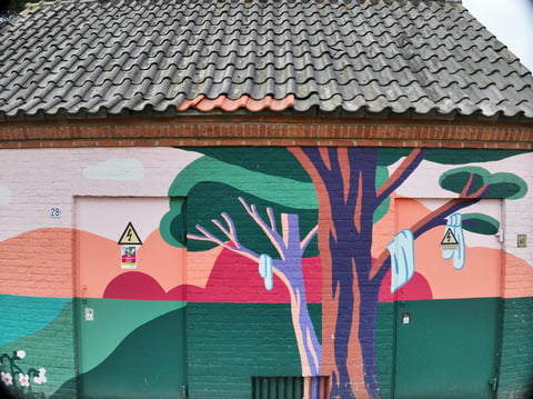 Colorful street mural on brick building wall featuring stylized tree with purple trunk and green foliage against orange background