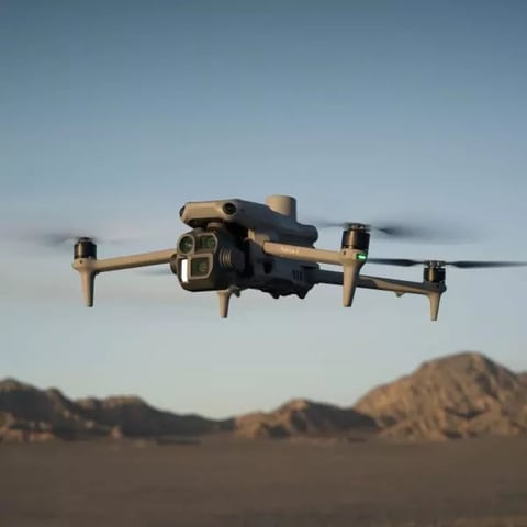 Tan and black compact drone hovering over desert landscape with mountains in the background