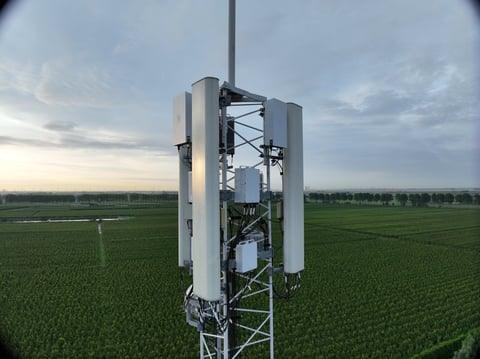 Cellular tower with white cylindrical antennas in rural farmland under overcast sky