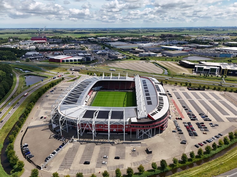 Aerial view of a modern football stadium with red seating, surrounded by parking lots and industrial facilities in a flat landscape